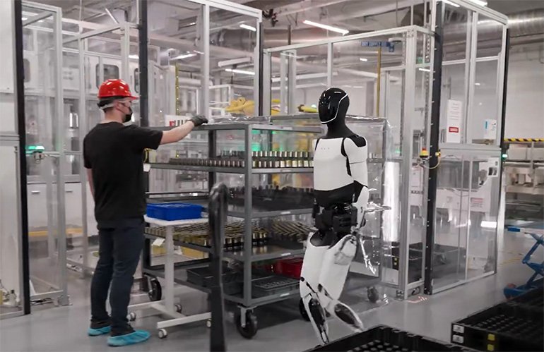 A Tesla Optimus humanoid robot walks through a factory with people. Predictable robot behavior requires priority-based control and a legal framework.